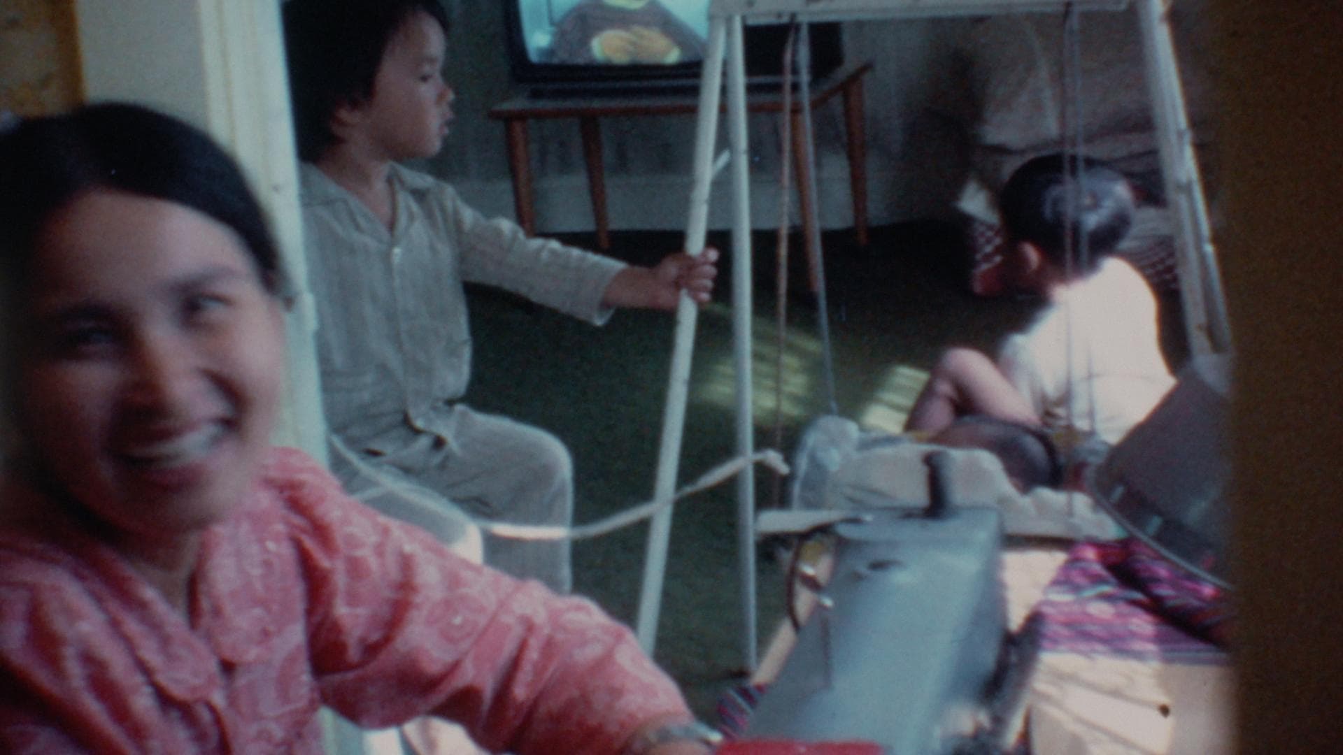 A film still from We Were the Scenery shows an Asian woman smiling as she is captured in motion in a door frame. Behind her, two Asian children rest. One child looks at the other, who is lying on a small swing and focused on a TV screen.