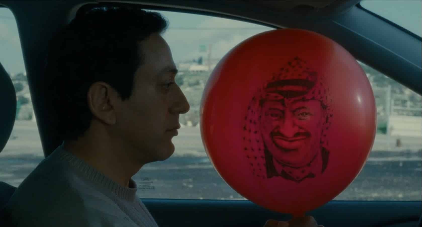 A film still from Divine Intervention shows a close up of a man in car, he has a somber expression and is holding a big red ballon with a person's face printed on it in black. The person on the ballon has a scarf on their head and is smiling menacingly.