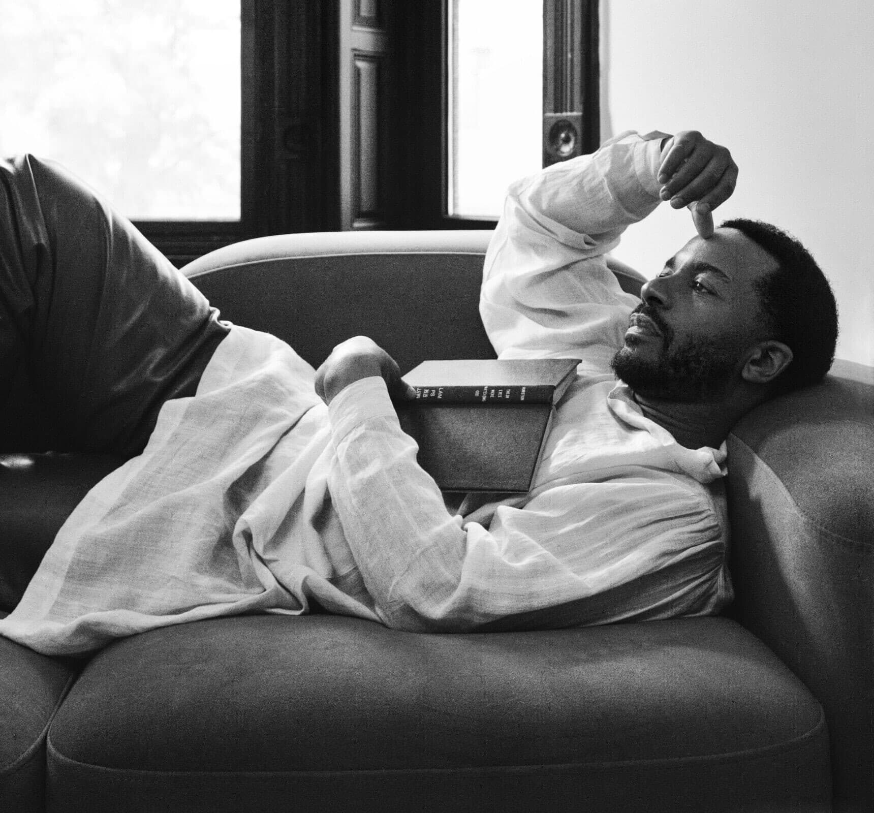 André Holland reclines on a couch, looking pensively into the distance. His arm is above his head, and a sliver of a book can be seen resting on his chest. The photo is in black and white.