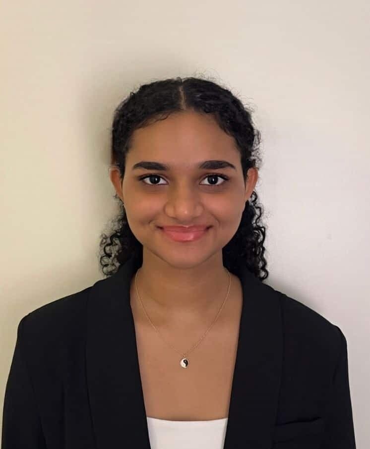A headshot of Ritikaa. They are a women of color, they have on a black blazer and a silver necklace. They are smiling at the camera.