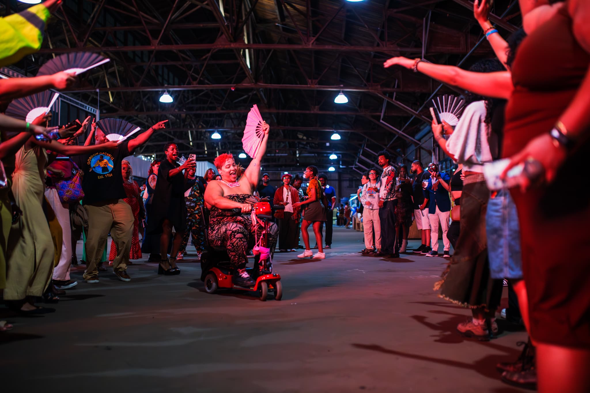 A photo from the 2025 BlackStar Film Festival opening party. A spirit tunnel has opened on the dance floor, a person in a wheelchair is going through it, they are holding a paper fan in the air. Those on the sides have paper fans as well, raised in celebration.