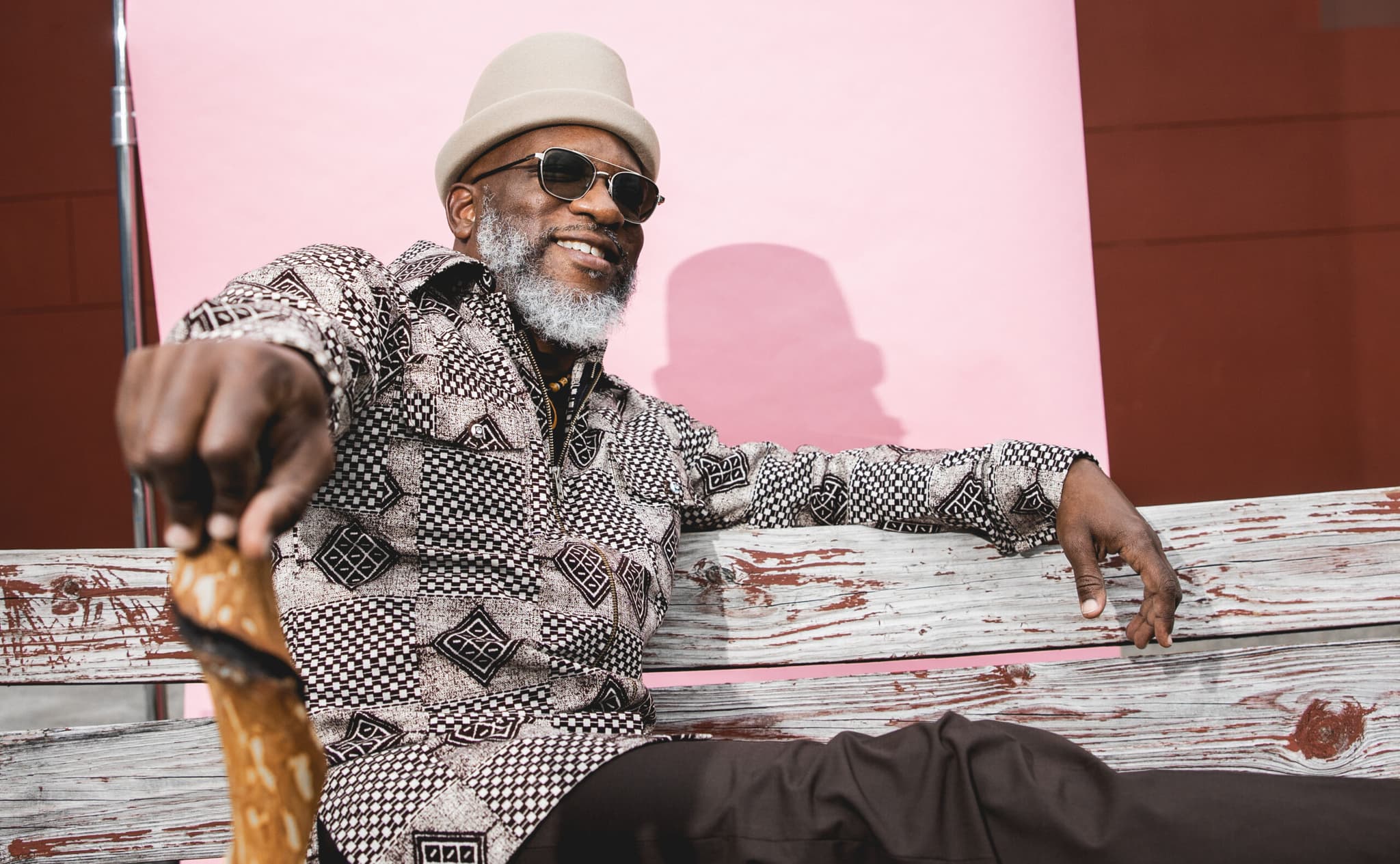 A headshot of Orrin. He is a Black man, he is wearing a white hat and sunglasses, he is sitting on a wooden bench.
