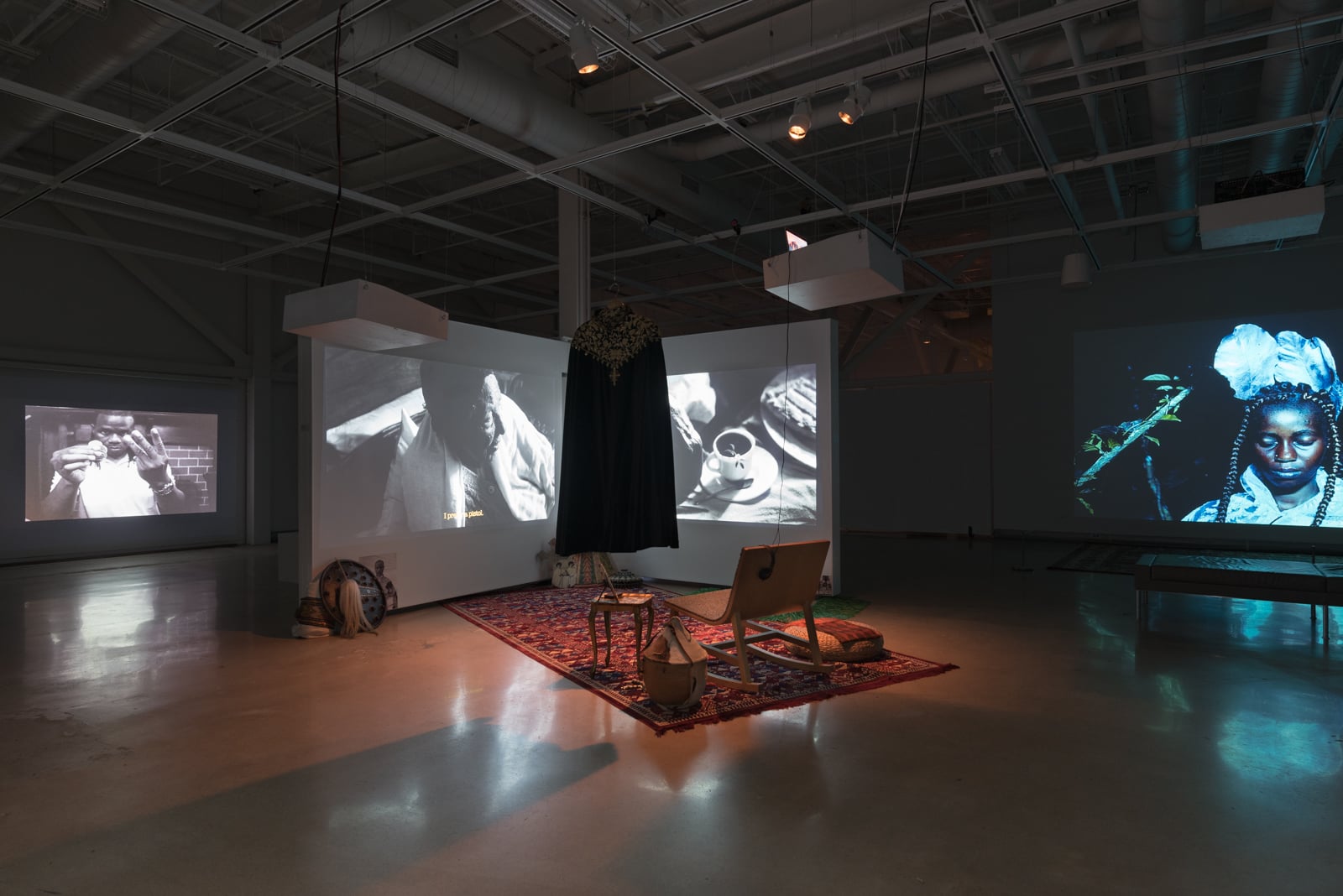 A photo of a gallery exhibition featuring multiple projected videos on white screens, plus a carpeted viewing area in the middle of an otherwise dark room.