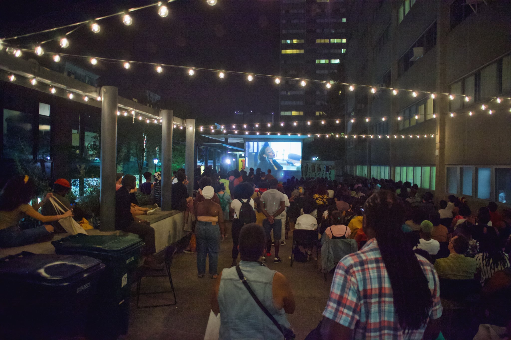 A group of people have their backs turned to the camera as they attend an outdoor film screening, standing under multiple strings of light at night.