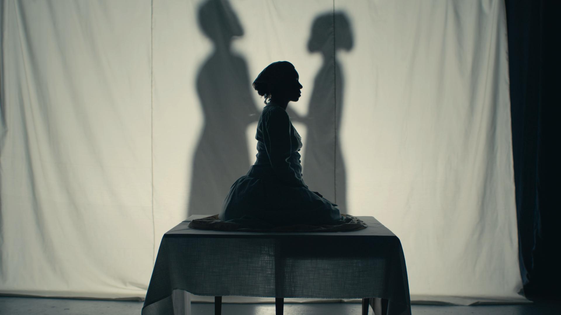 A still from Bone Black: Midwives vs. the South. It shows a silhouetted woman sitting on a pedestal, there are shadows of people behind her.