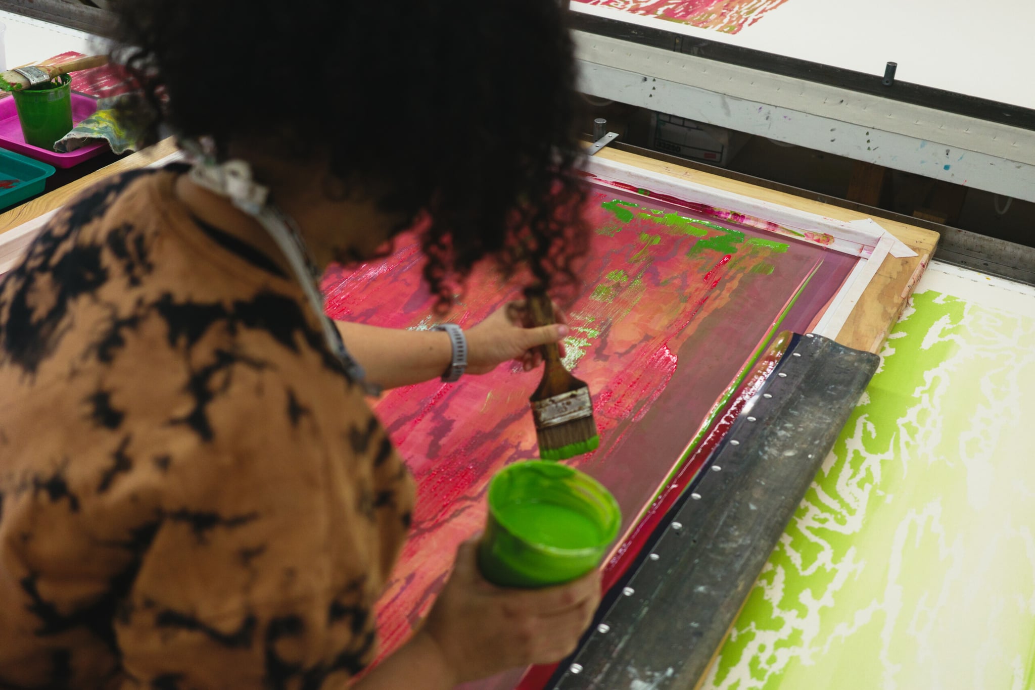 A photo taken over the shoulder of an artists with brown curly hair painting a canvas while holding a cup of bright green paint.