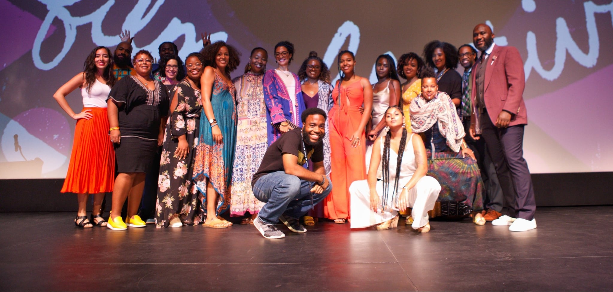 A group of Black, Brown, and Indigenous people gather on a stage in front of a large screen (which displays the words film and festival). They are all smiling and a few in the front are posing, leaning down the make room for others.