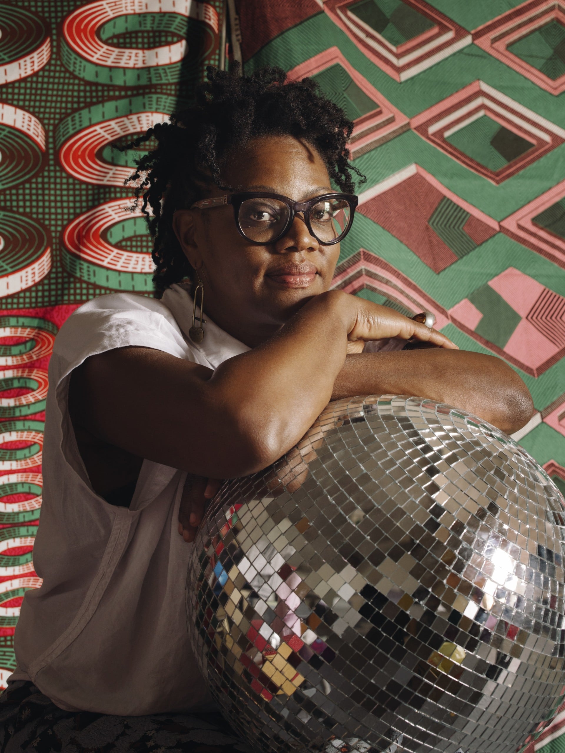 A photo of Cauleen Smith, who is a Black person wearing black-framed glasses, arms-crossed over a disco ball, in front of a colorful print.
