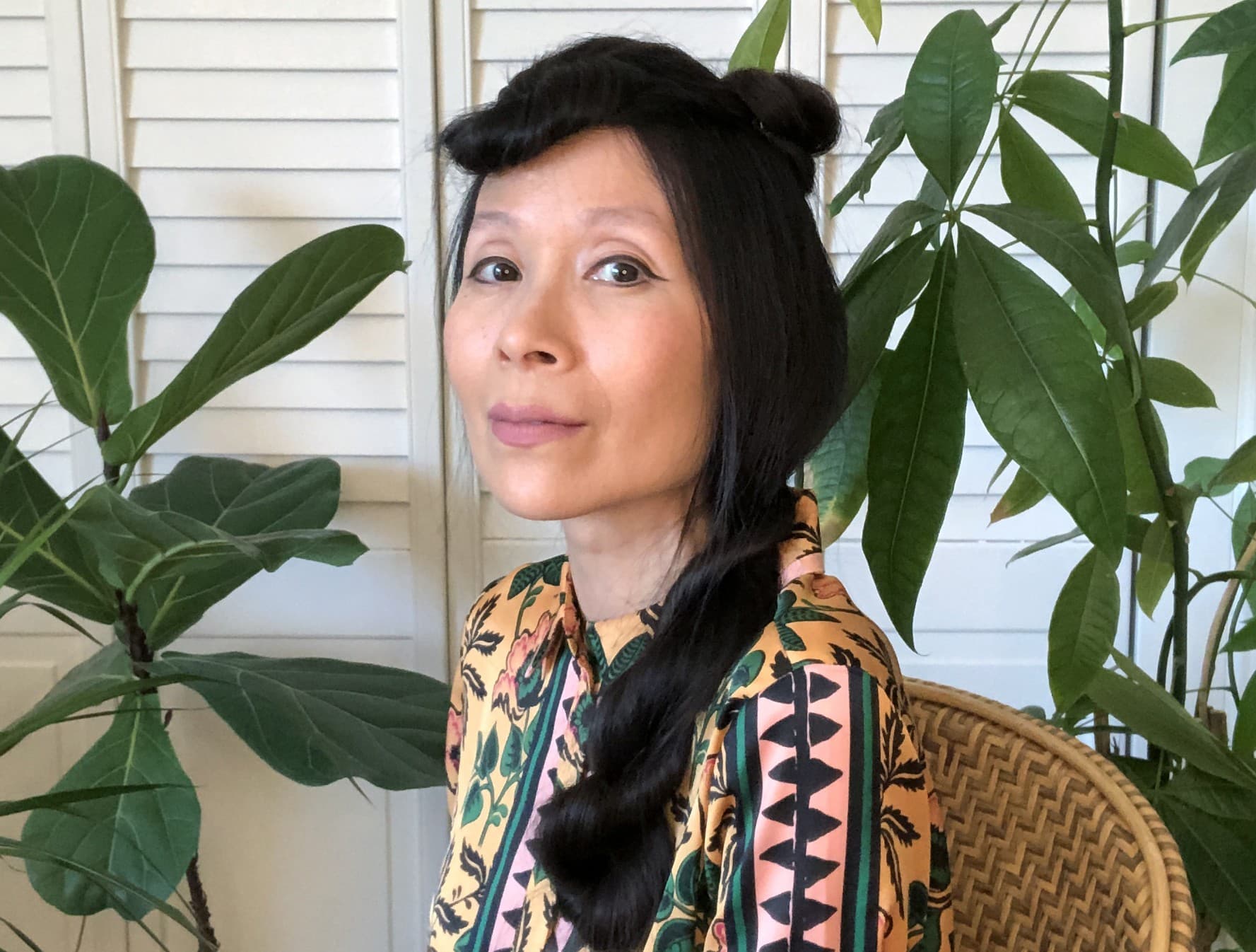 A photo of Astria Suparak. She is sitting in a wicker chair and there are plants behind her. She is wearing a gray skirt and a patterned top. She is smiling and looking at the camera.