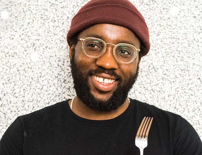 A headshot of Tunde Wey, he is wearing a burgundy beanie, a black shirt and has on clear glasses. He is holding up a fork, smiling, and looking slightly towards the left.