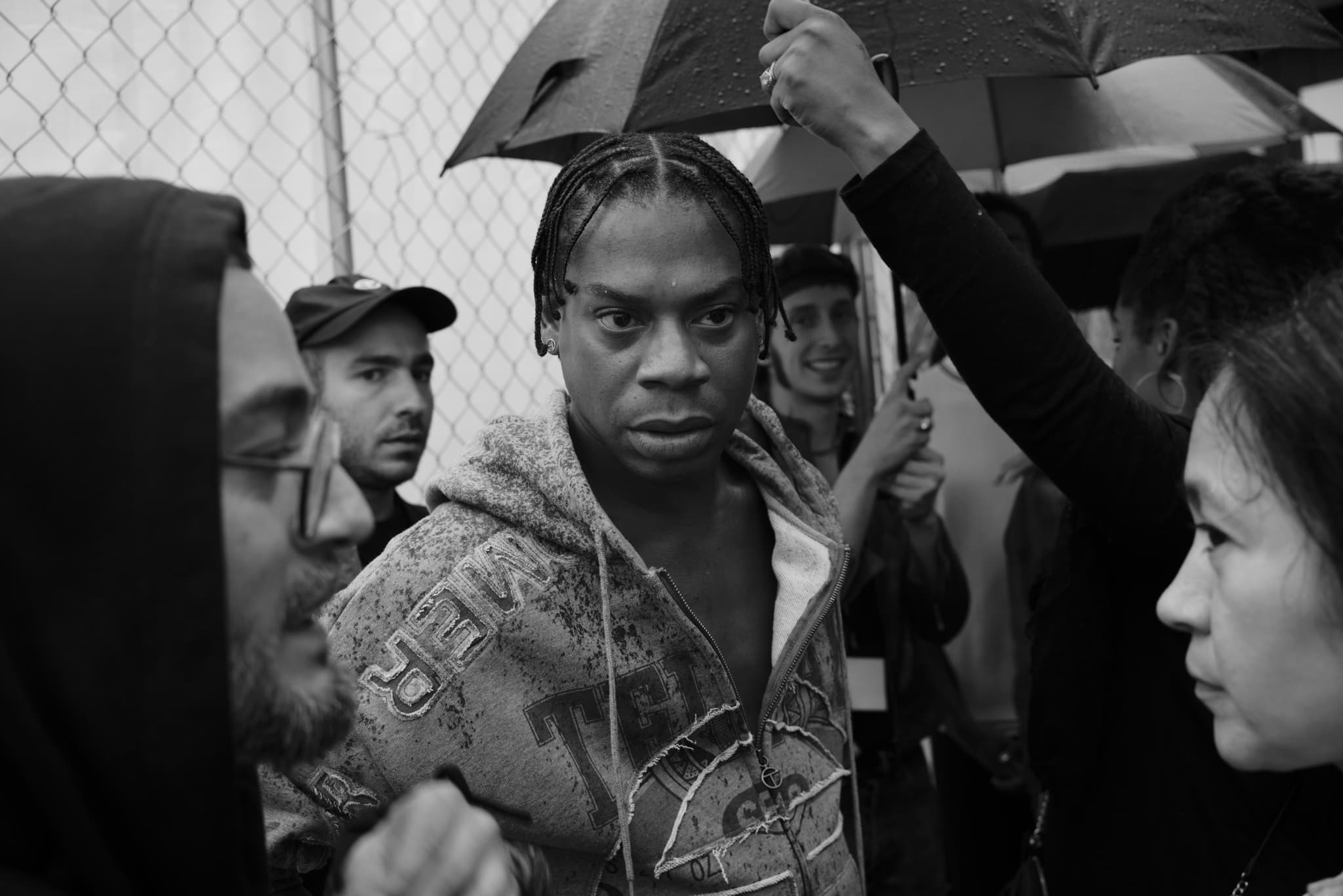 A black-and-white photo of Avena, Babak, and Telfar. They are outside and it's raining, someone is holding an umbrella in the background. The man in the middle, presumably Telfar, has short braids and is wearing a sweatshirt. He has a serious expression.