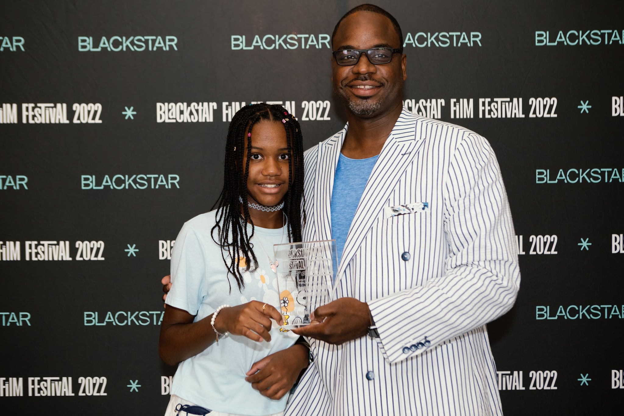A photo of Bria and Bilal Motley, a Black father-daughter duo, they're standing in-front of a step-and-repeat that says "BlackStar Film Festival 2022." They are holding an award.
