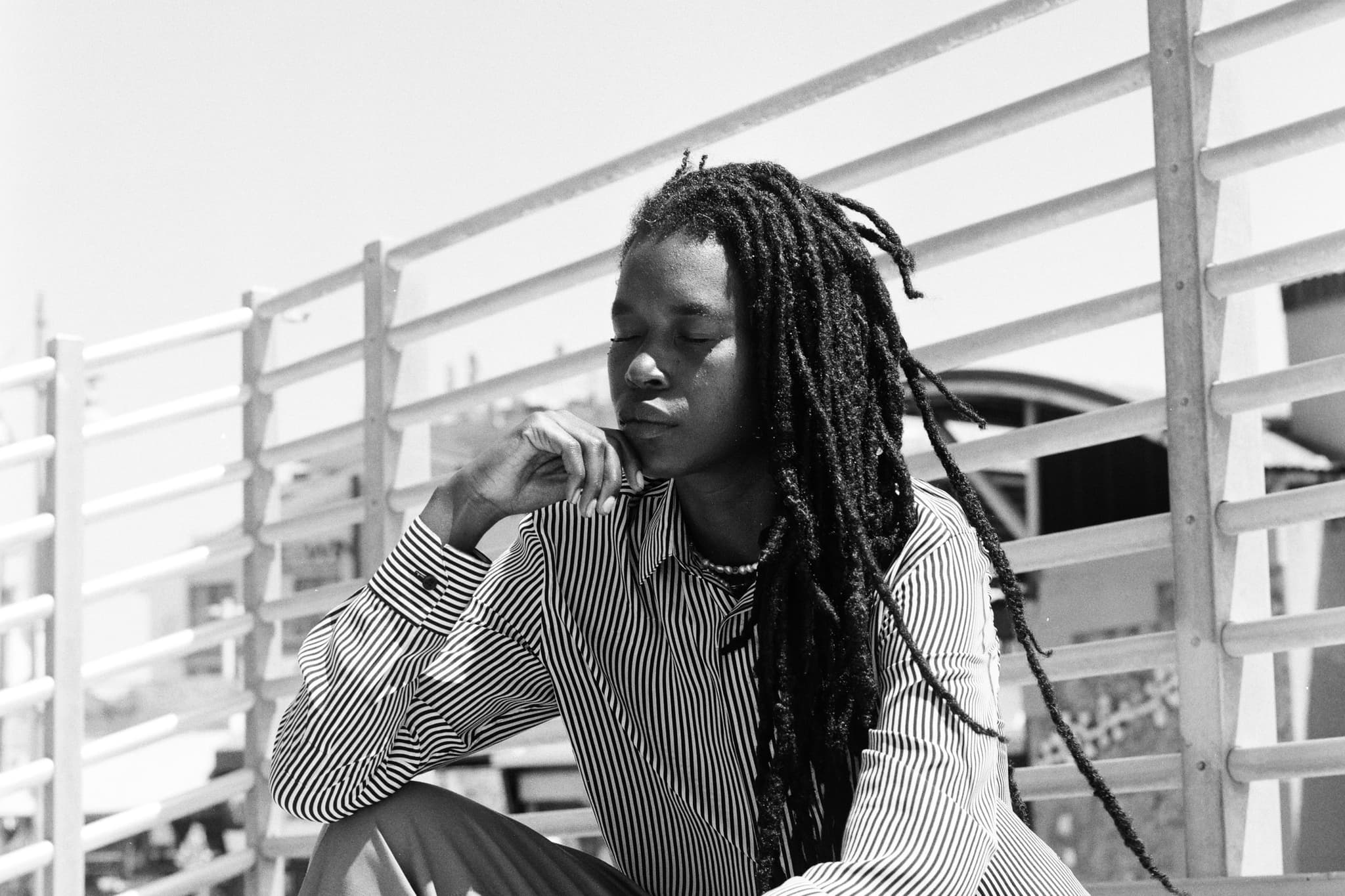 A black and white photo of Camae Ayewa/Moor Mother. They are sitting in front of a fence. Their eyes are closed. They have long locs that fall around their head. They have on a pin-stripped shirt.