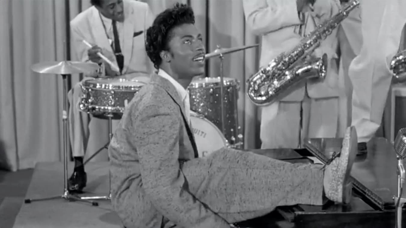 A black and white photo of Little Richard. A Black man stands on stage with one leg stretched into the air. He is wearing a suit and a bee hive-esque hair style, and is standing in front of a band only partially visible.
