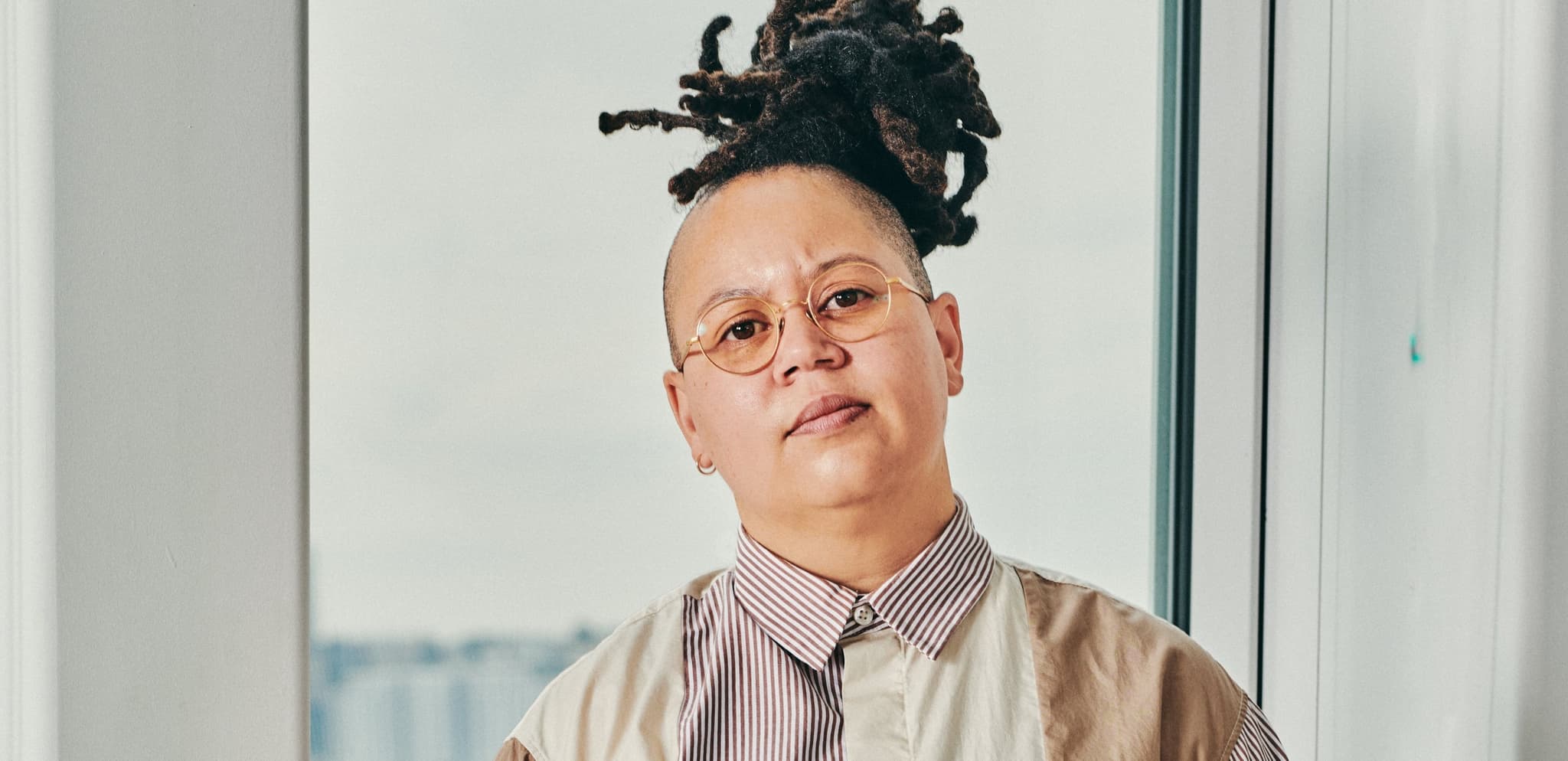 A headshot photo of Meg Onli. They have a light skin-tone. The sides of their head are shaven and they have long locs tied up into a bun. They are wearing a mutli-patterned button down shirt.