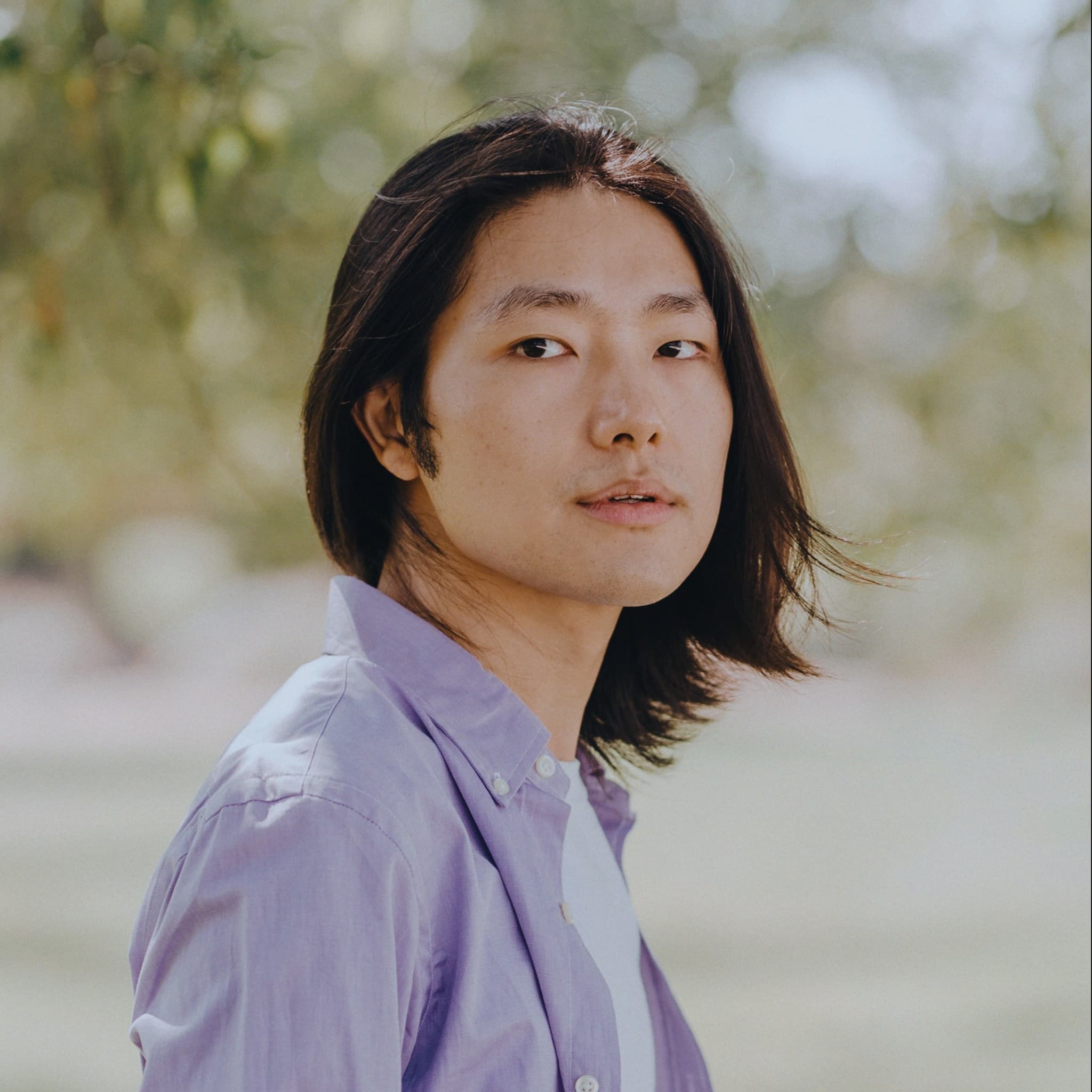 A picture of a non-binary person named Zhou Hao. He has long, straight black hair that frames his face and slightly falls towards his shoulders. He is wearing a light purple shirt over a white top. The background is softly blurred, comprising natural elements such as trees and greenery. His expression is neutral, yet slightly contemplative, with his gaze directed slightly off-camera.