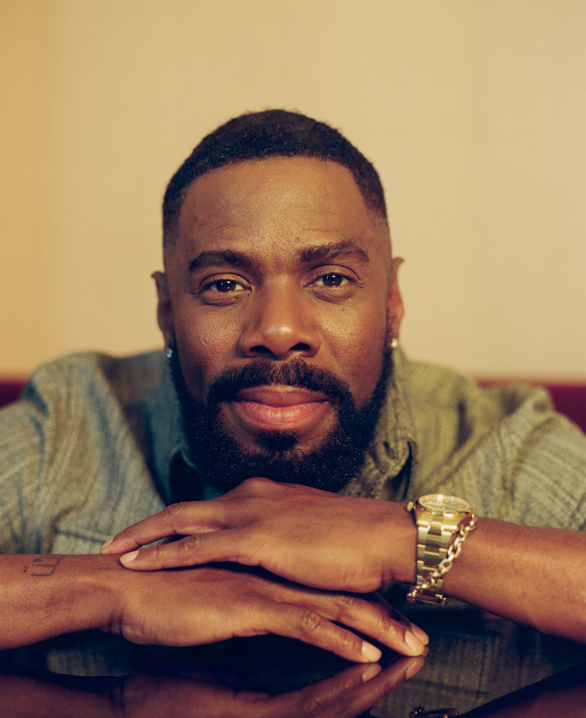 A close-up photo of Colman Domingo, who has his hands folded in front of him on a table, and is resting his chin on those hands. He wears a gold watch and a green top, and seems to be on the verge of a smile.