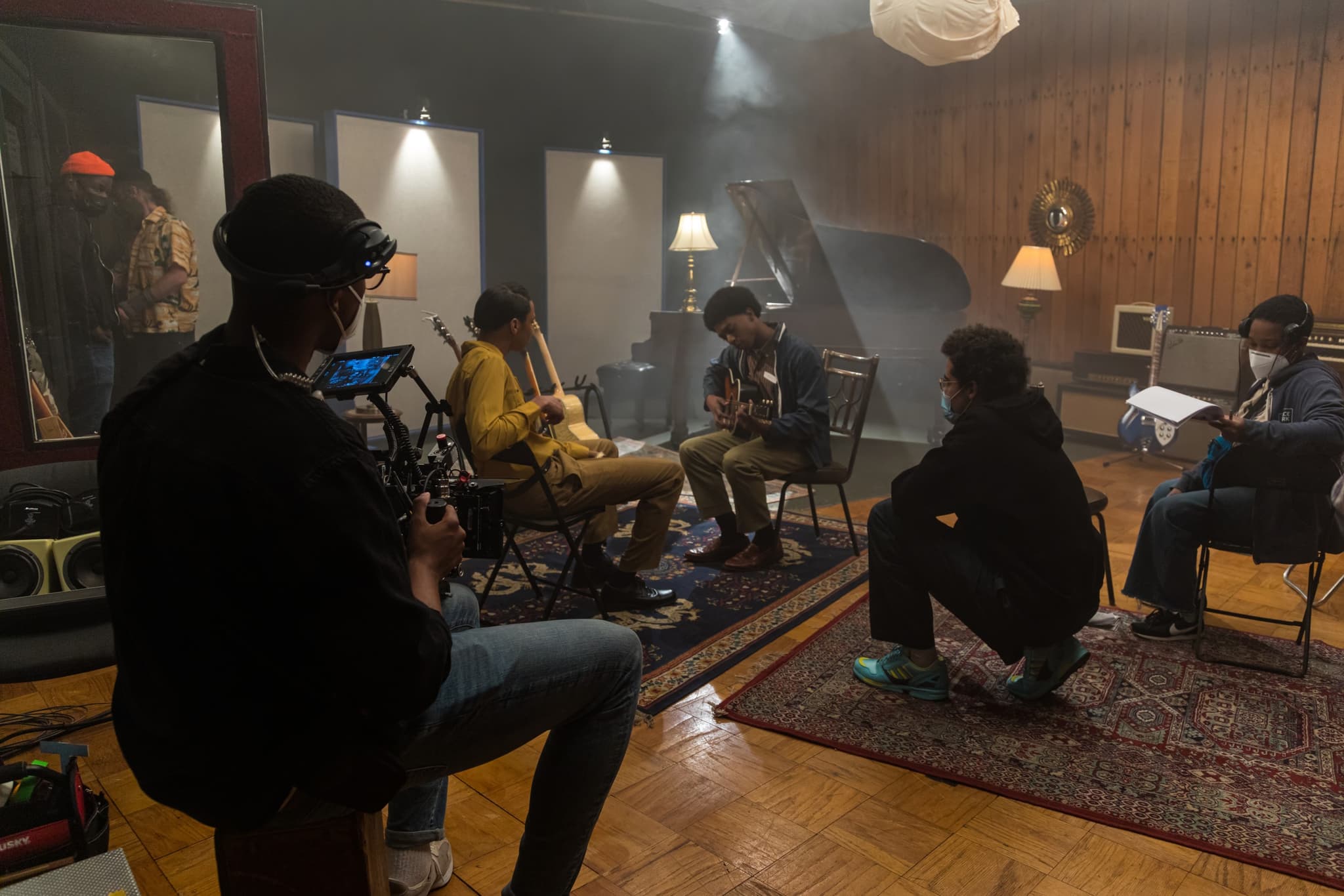 A photo on set of the 2022 BlackStar Philadelphia Filmmaker Lab. It shows the cast in the middle of scene, a young boy is holding a guitar. The director kneels beside him, presumably providing direction. There is a crew member in the foreground with a camera.