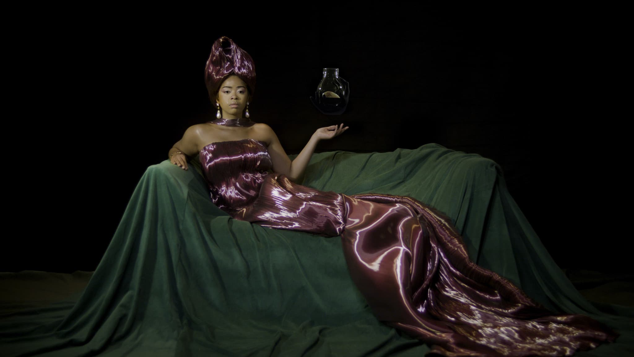 A still from All That's Left shows a Black woman dressed in a soft purple silk gown and a wine glass sitting on some kind of display.