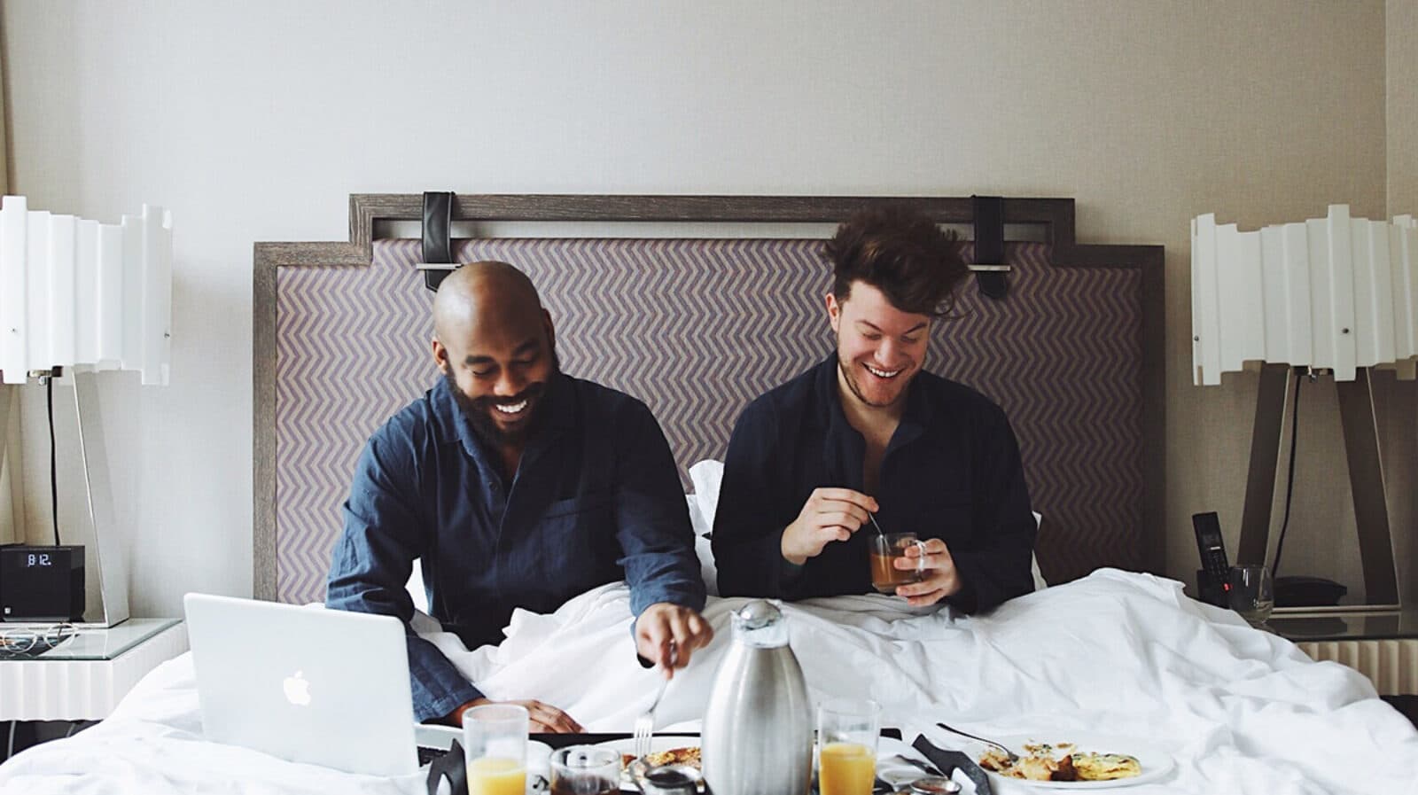Two people, one Black and one white, are having breakfast in bed at a hotel.
