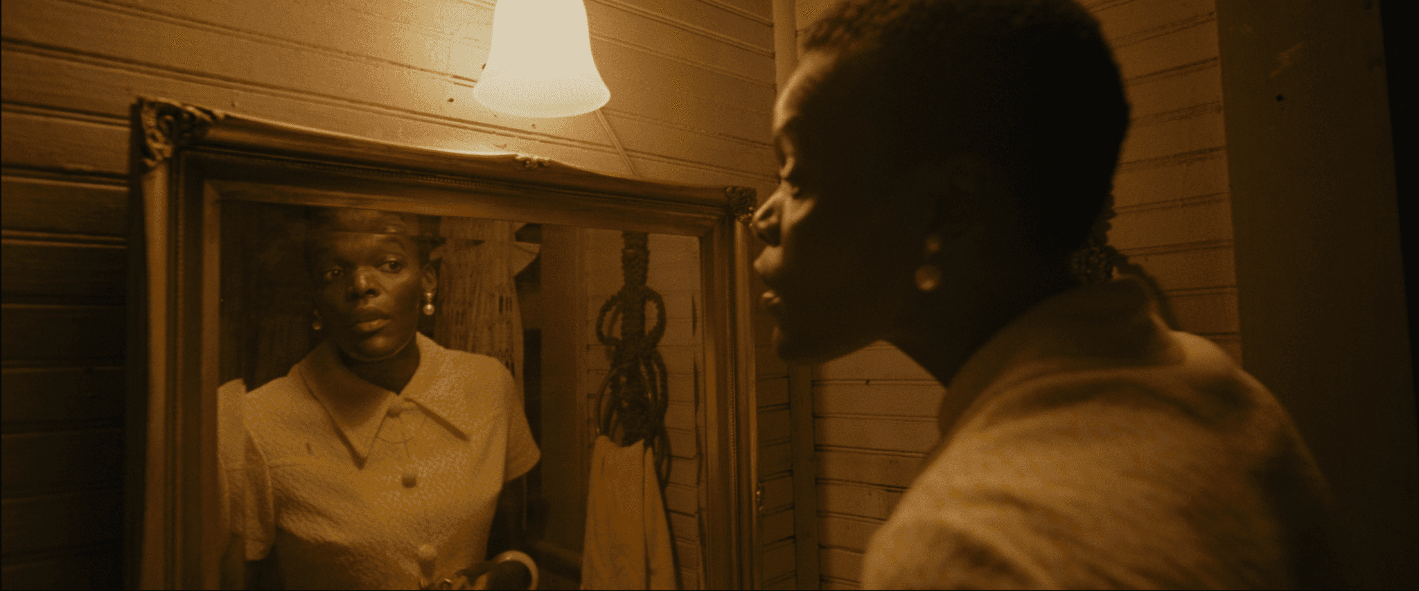 A woman wearing red lipstick and a white dress stands in a bathroom. She looks at herself in the mirror. A light above the mirror emits a yellow glow.