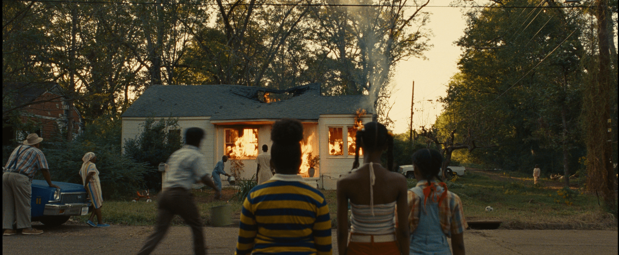 In a wooded neighborhood, a white house is engulfed in flames. Near the house, a person stands in white, their back to the camera, watching the house burn. On the left, a man in blue coveralls throws a bucket of water on the fire. Two other people watch off to the side. Another man rushes from the street towards the fire. On the street, furthest away, three young girls dressed in colorful outfits stand still in front of the fire, only their backs are visible.