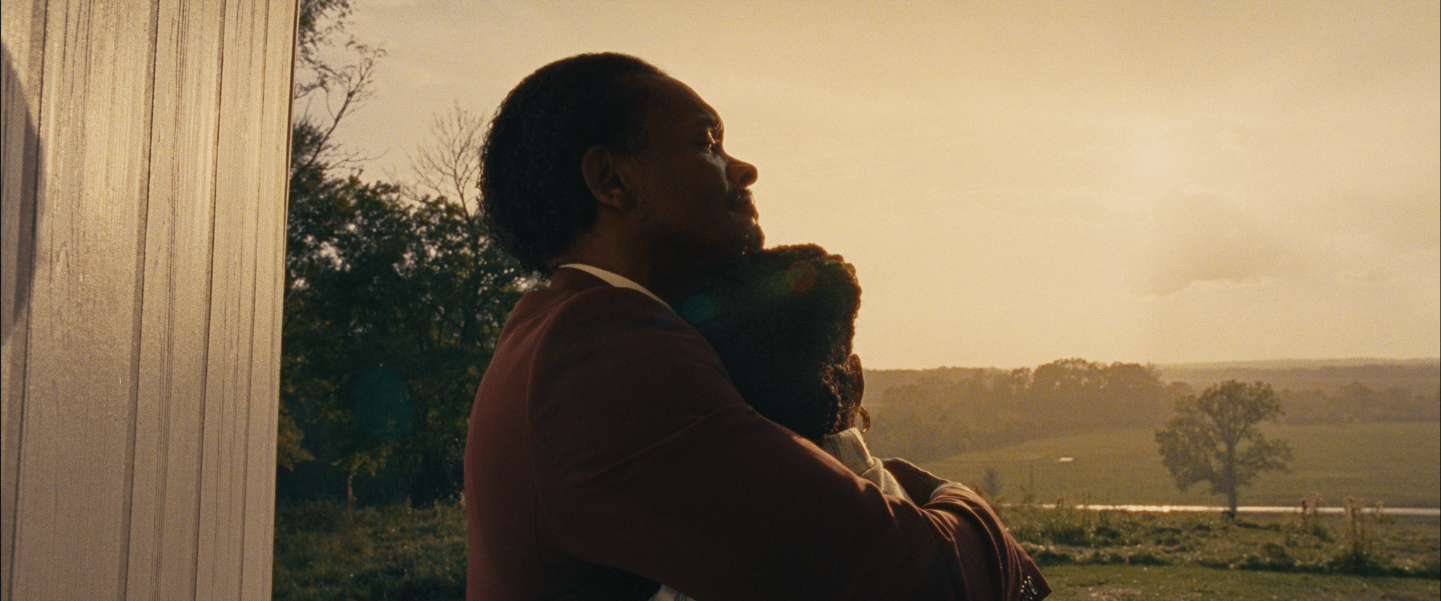 A man and child stand in front of a window. The window overlooks an open green grassy field with trees scattered throughout. The sun emits a yellow hazy glow over the field. In the foreground, the man and child hold one another. Only the man’s profile is visible while the young girl faces away from the camera.