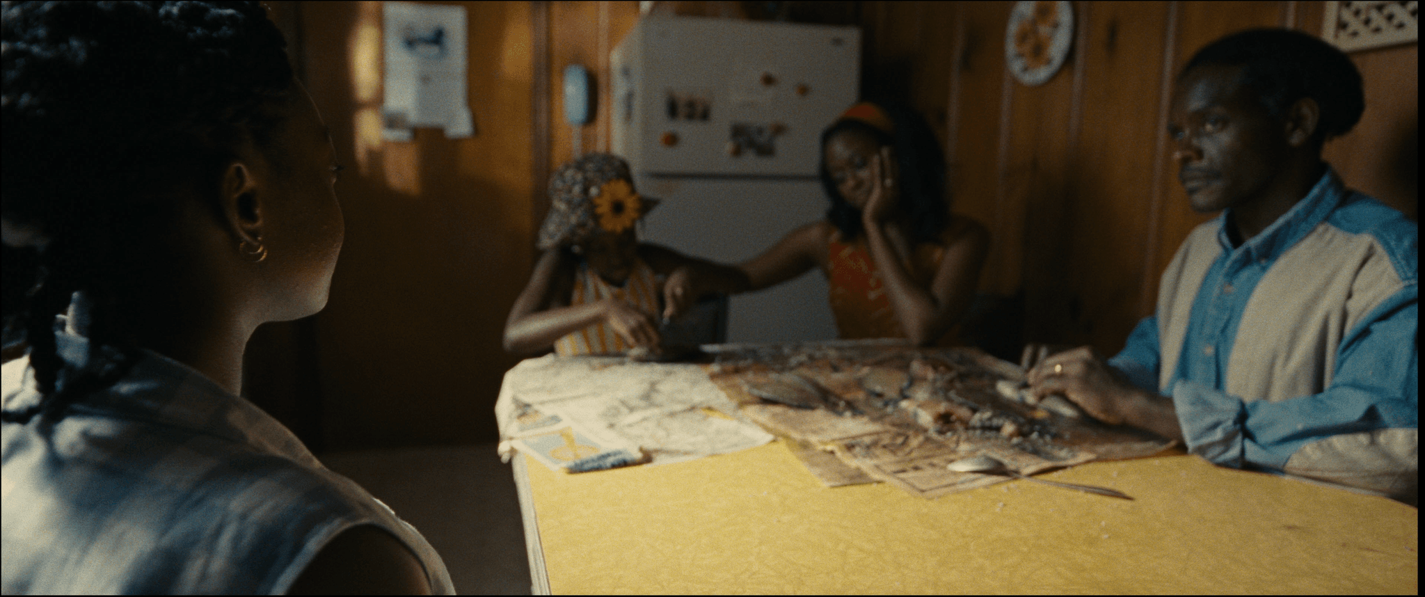 Four people sit around a yellow kitchen table. On the table, there is a half gutted fish with some of its parts strewn over a piece of newspaper.