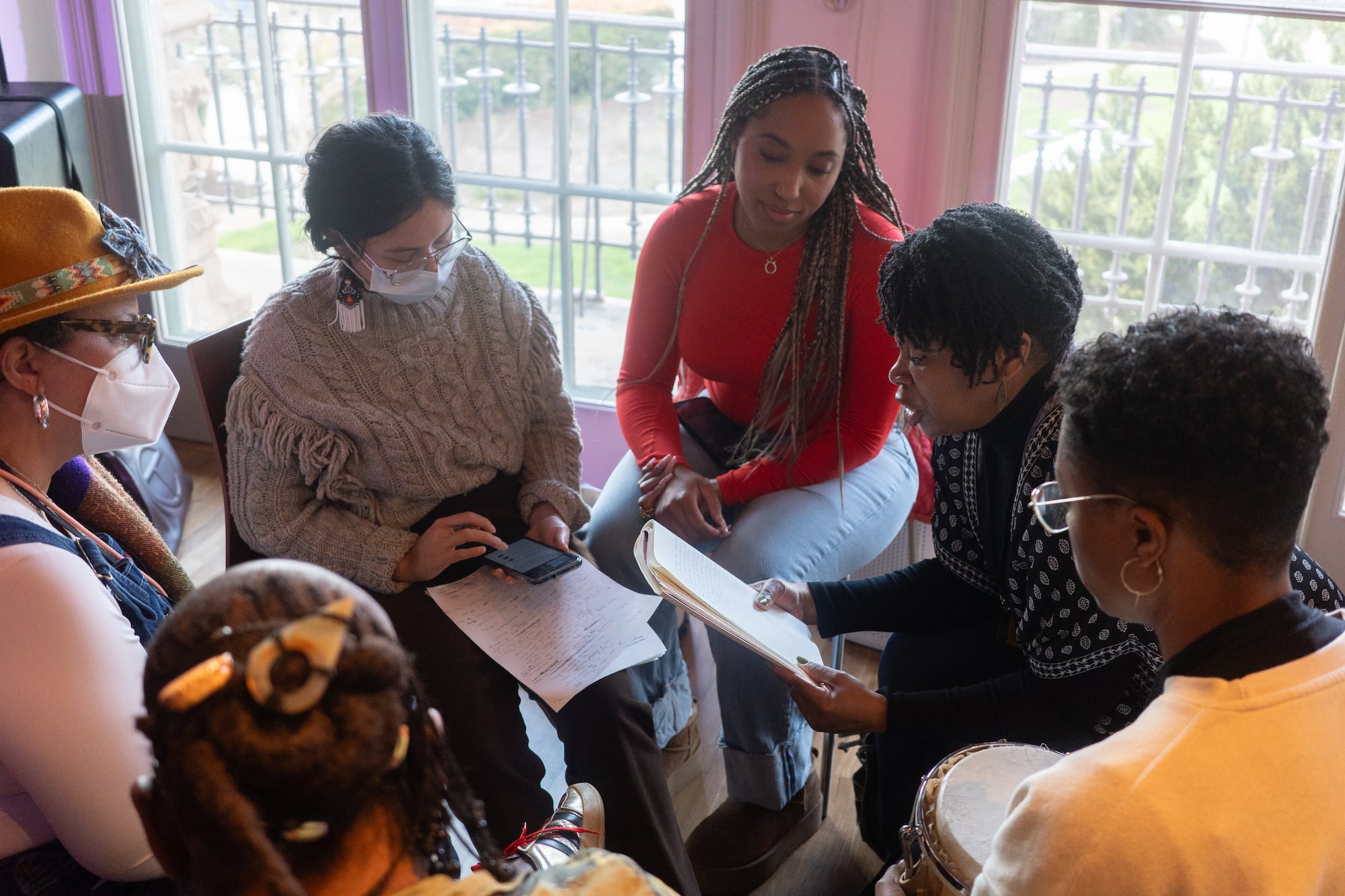 A photo of a group of people sitting together, one is looking at a journal and reading from it to the ground. There others appear to be listening intently.
