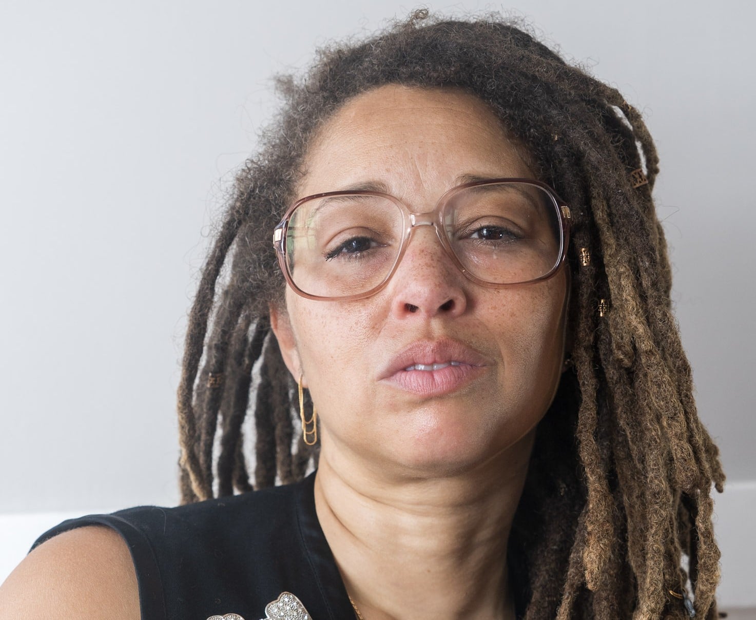 Maria Judice is a Black person. They have long locs. They are wearing glasses and are staring ahead at the camera.