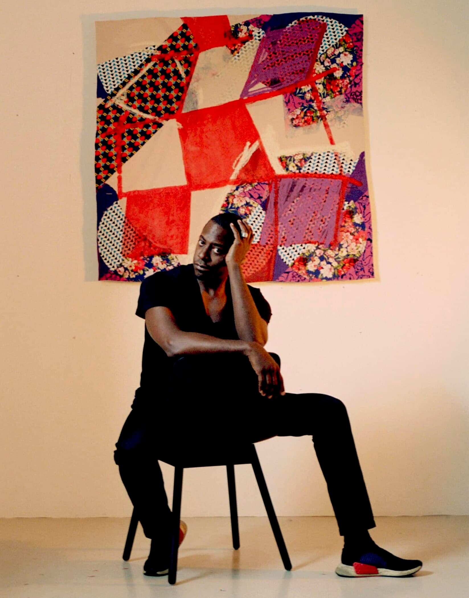 Sanford Biggers sits backwards on a chair while wearing all black with a quilted artwork on the wall behind him. The work is a patchwork of differently patterned fabrics and red quadrilaterals painted on top.