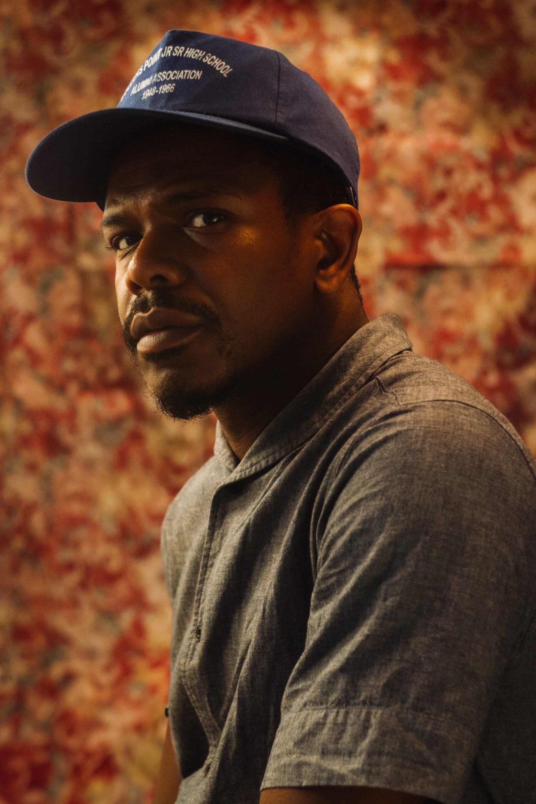 A portrait of Lendl Tellington. He is a Black man. He is wearing a navy blue cap and a gray button-down shirt. He is looking at the camera with a contemplative expression.