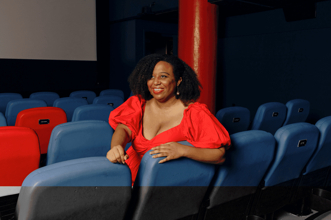 A photo of Maya S. Cade. She is a Black women. She is sitting in a movie theater. She is wearing a bright red dress. She has curly, long black hair.