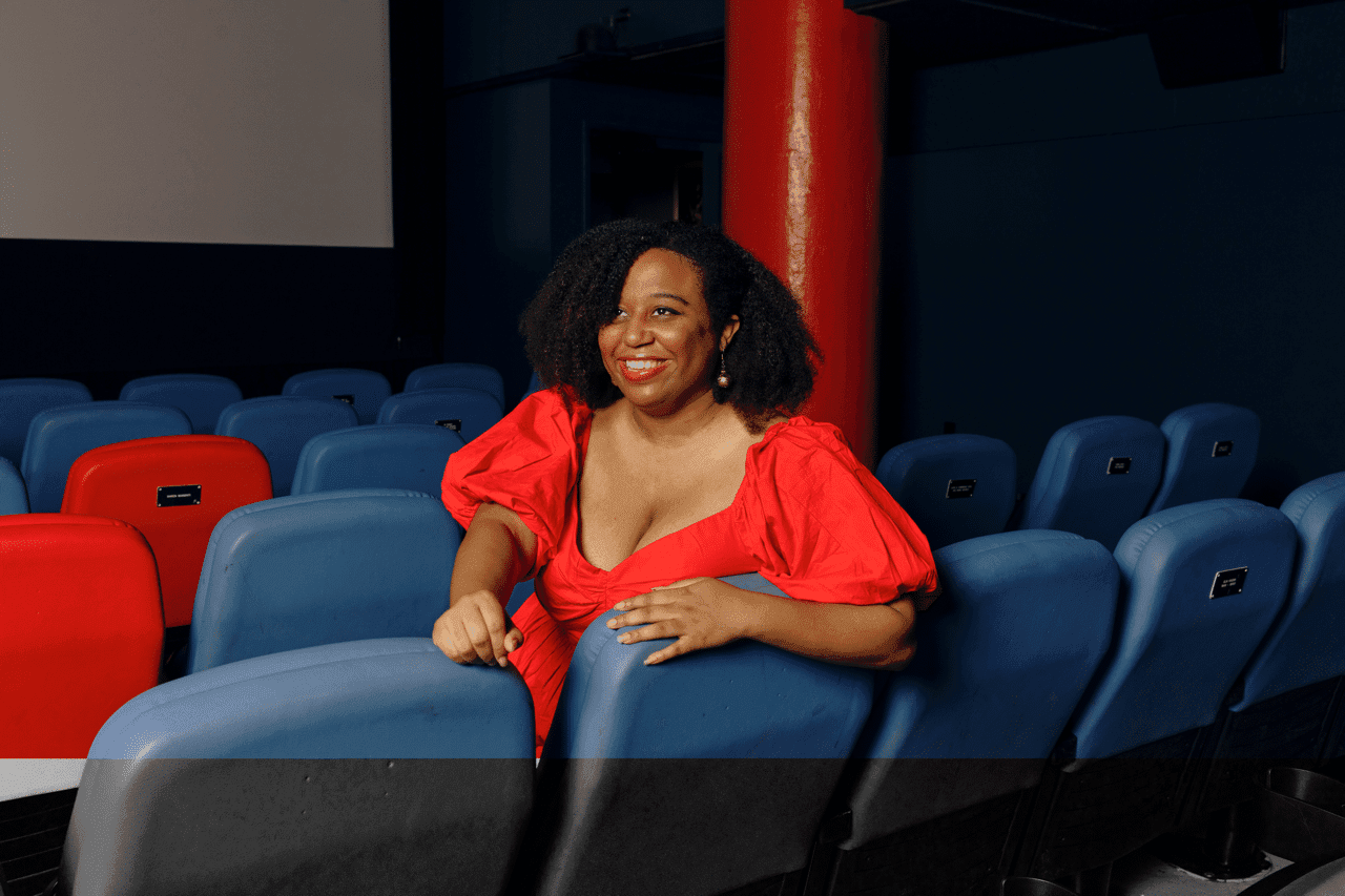 A photo of Maya S. Cade. She is a Black women. She is sitting in a movie theater. She is wearing a bright red dress. She has curly, long black hair.