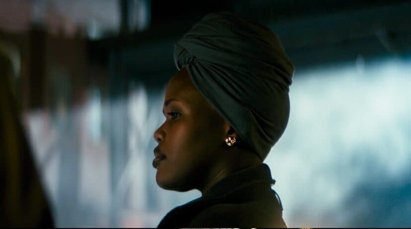 A still from Adamstown shows the side profile of a Black woman staring at something off screen. She is wearing a green scarf tied in a knot on her head and gold earrings against a blurred out background.